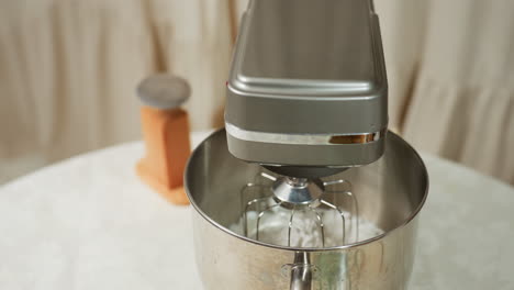 electric mixer placed on kitchen table with white dough inside stainless bowl, surrounded by additional kitchen tools including wooden base stand and spatula tips, ready for baking