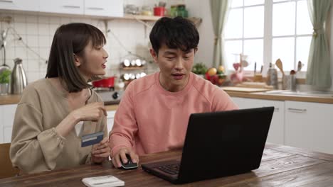 happy asian married couple enjoying shopping online with credit card and laptop. the man is keying in the number to make payment as the woman is reading to him