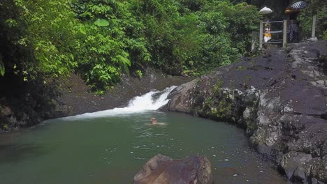 beautiful blonde woman swims in waterfall pool in indonesian jungle