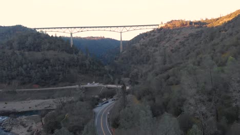 drone flying high up over the american river with a view of the famous foresthill bridge in the auburn, california during sunset - surrounded by green trees and mountains