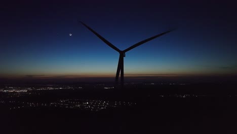 a wind turbine stands against a twilight sky, city lights twinkling below