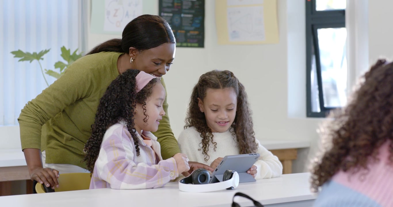 Insegnante afroamericana che si avvicina alla scrivania, aiutando due bambine che usano un tablet e delle cuffie