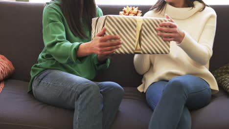 midsection of biracial mother and adult daughter exchanging christmas gift on couch, slow motion