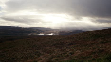 Fast-winds-blow-grass-on-the-side-of-a-Scottish-mountain-as-shafts-of-light-break-through-dark,-moody-clouds-in-the-background-to-highlight-fresh-water-lochs-amongst-the-mountains