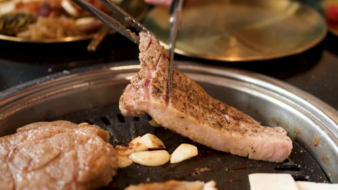 Hand cuts pork belly with tongs and scissors on tabletop Korean barbecue grill, warm lighting