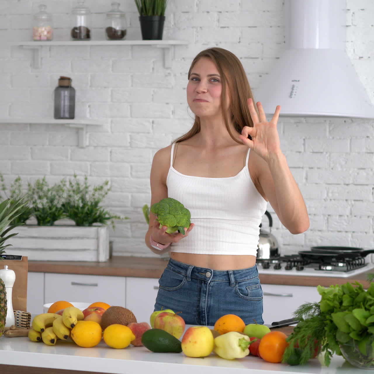 Healthy woman with sporty body in the kitchen. Attractive girl holding broccoli and shows her biceps on camera. Healthy food on the kitchen table at home.