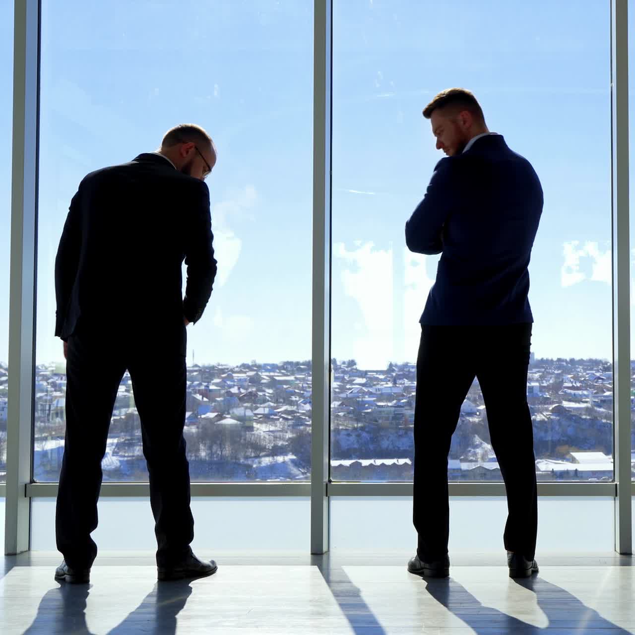 Elegant entrepreneurs meeting indoors. Businessmen in dark suits having a conversation near the large office window and looking on the city landscape. Back view