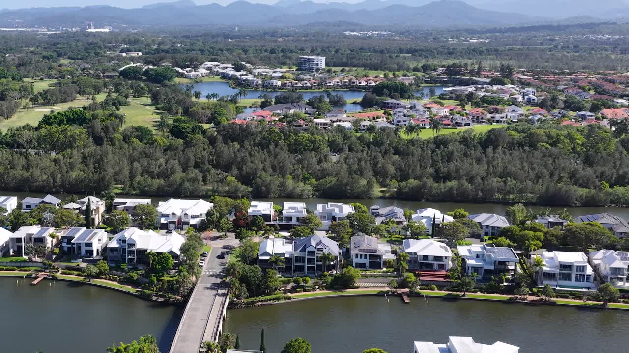 Barrido con dron de una zona residencial soleada junto a un lago con casas modernas, un puente, vegetación y colinas a lo lejos