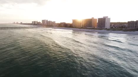 Drone-Volando-Hacia-La-Playa-Al-Atardecer-Cerca-De-Un-Muelle