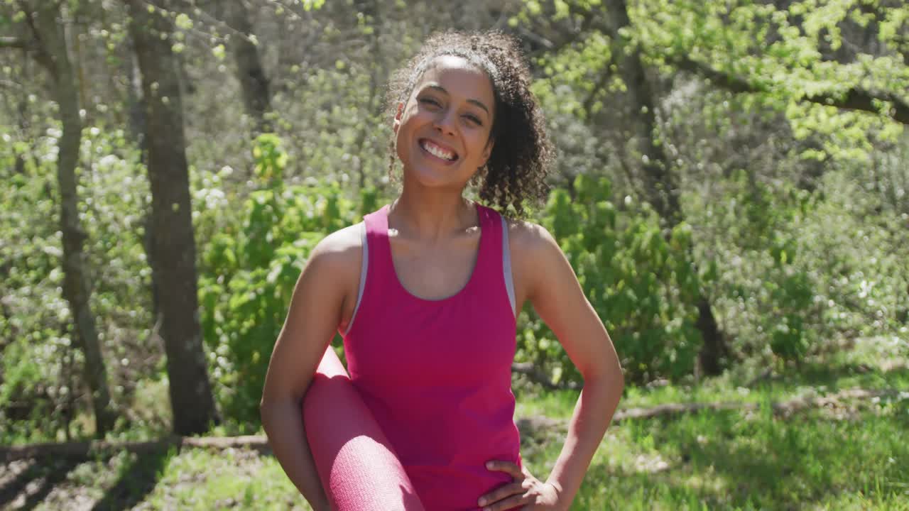 retrato de una mujer biracial sonriente con ropa deportiva en el bosque con colchoneta de yoga