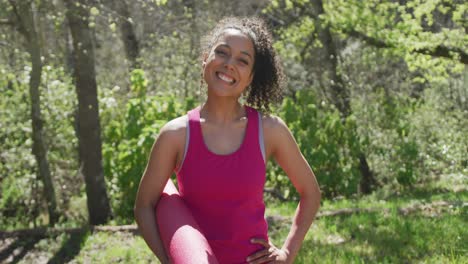 retrato de mulher biracial sorridente vestindo roupas esportivas na floresta com esteira de ioga