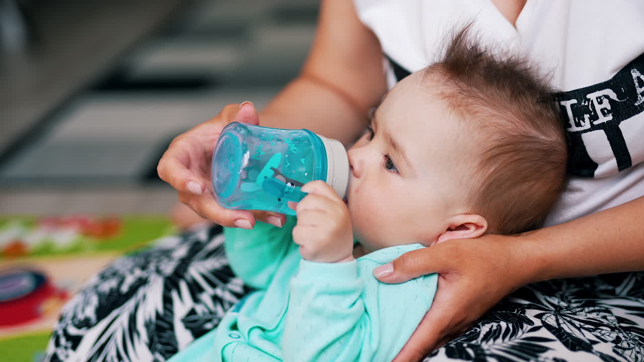 Mommy is giving a bottle with water for her baby. Cute child doesn't want to let go the bottle. Close up.