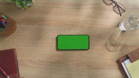 top down view of a laptop computer with mock up green screen chromakey display on a wooden office desk next to notebook with pens, glasses, and a glass of water. slow zoom out, close up