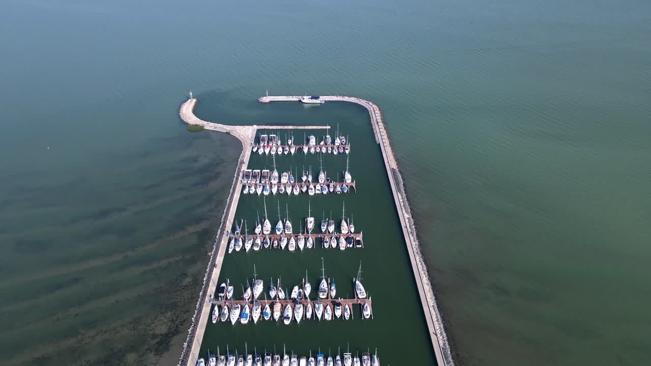 An aerial footage of a marina on a serene lake, with rows of boats and yachts docked within a breakwater.