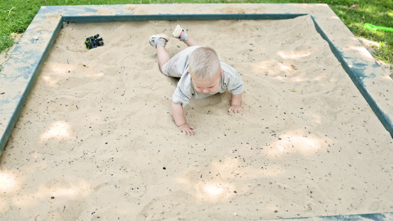 Un bambino in maglietta e pantaloncini giace in una sabbiera. Il bambino piccolo gioca con la sabbia in un parco giochi. Il bambino si diverte in una sabbiera. Il bambino passeggia all'aperto.