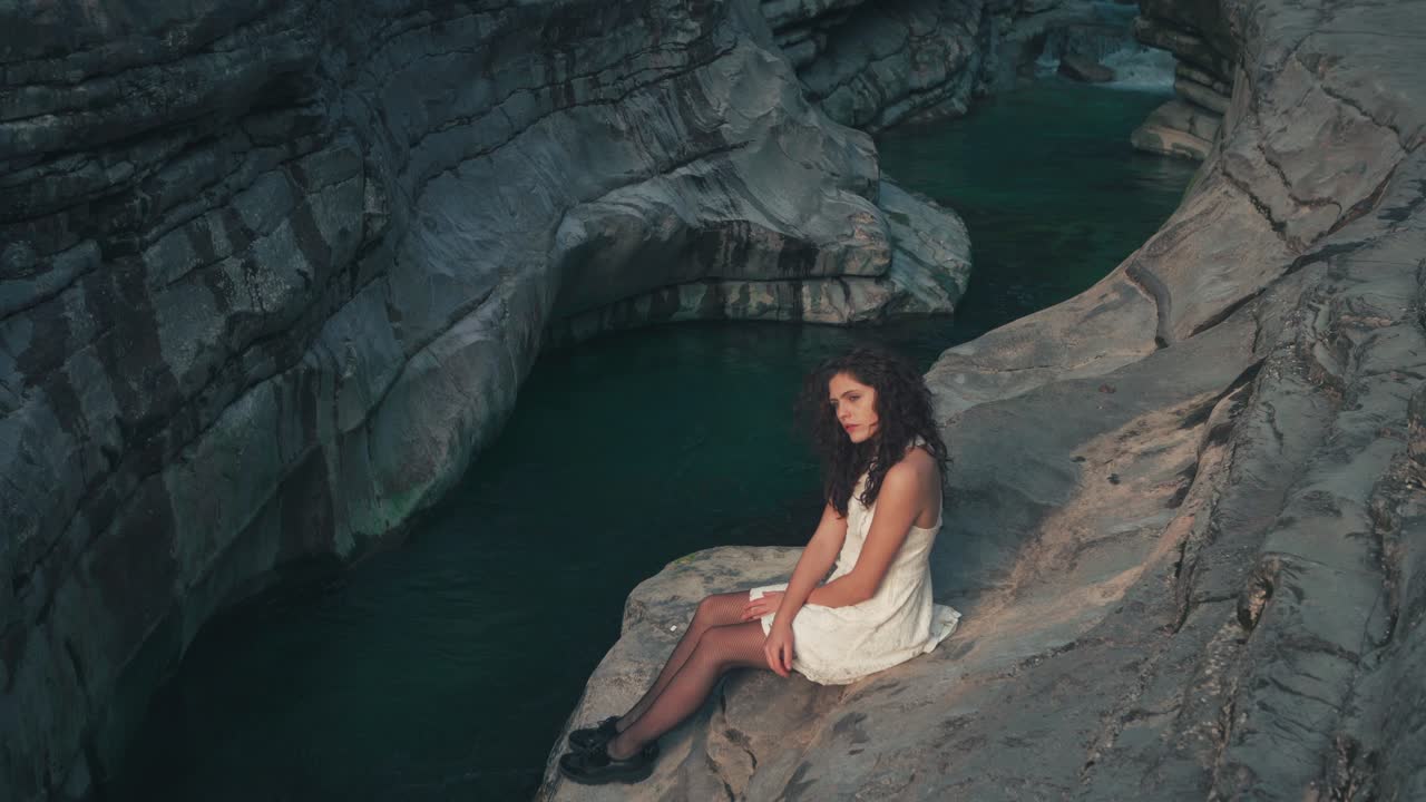 4K Cinemagraph - seamless video loop of a beautiful young brunette model woman in a white dress sitting at a gorge by a blue mountain river. The water is rushing through naturally formed rocks.