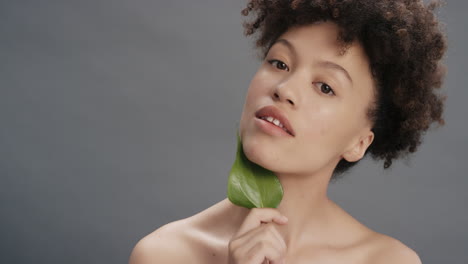 Retrato-Hermosa-Mujer-De-Raza-Mixta-Sonriendo-Acariciando-La-Cara-Con-Hojas-Tocando-Una-Tez-De-Piel-Suave-Y-Saludable-Disfrutando-De-La-Belleza-Natural-Del-Cuidado-De-La-Piel-Sobre-Fondo-Gris