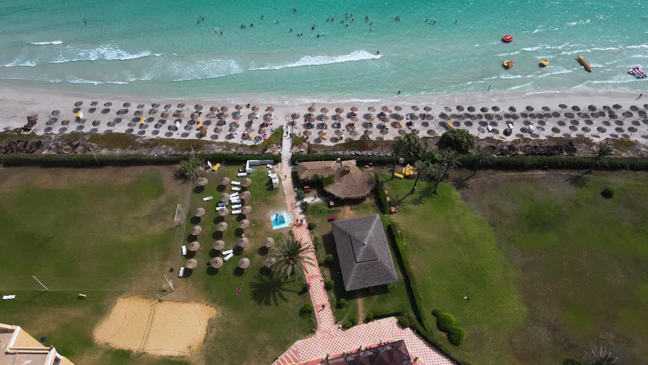 The image is an aerial view of a beach with crystal blue water of a Hotel at Mahdia, where people are enjoying various activities. A few surfboards can also be seen floating on the water.