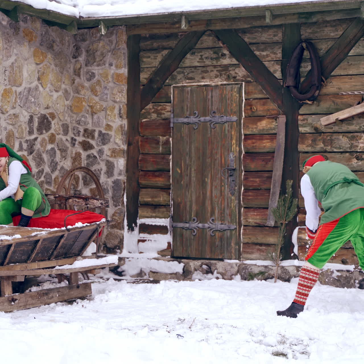 Joyful elves playing snowballs outdoors. Female elf sitting on wooden sleigh. Fairy elves in winter on the background of old wooden house. Christmas atmosphere.