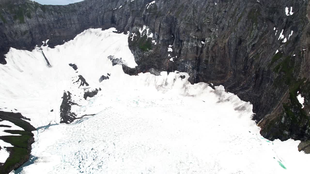 Tilt-up shot of a frozen lake covered in snow, with a glacier and towering rocky mountains in the background