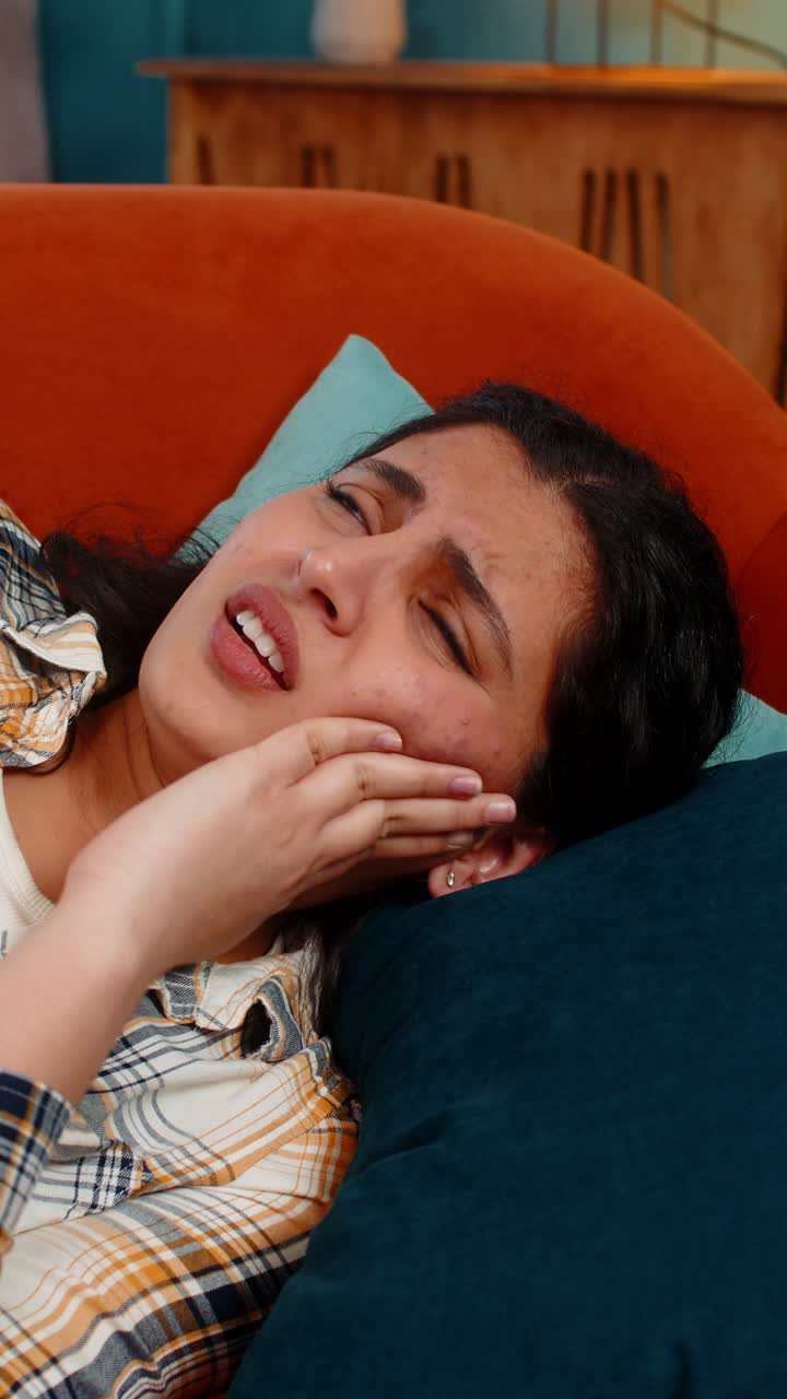 Indian woman holding cheek lying on sofa experiencing severe toothache pain discomfort distressed