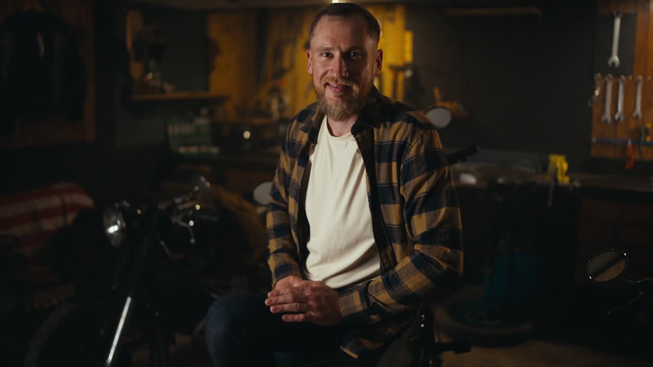 retrato de un feliz motociclista rubio con barba en una camisa a cuadros sonriendo y posando en su bicicleta en un garaje de taller