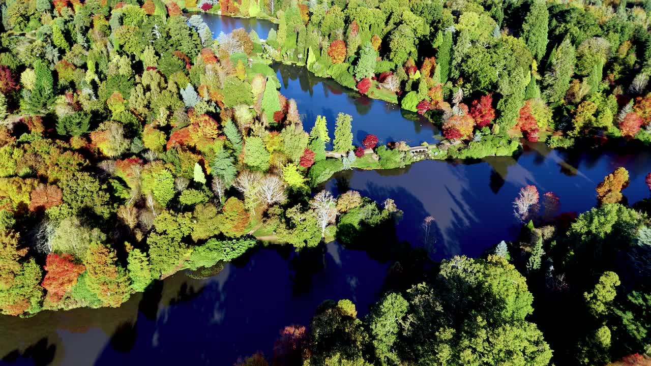 Aerial pan drone footage over autumn woodlands and shimmering lakes in Sussex, revealing a small bridge surrounded by vibrant foliage and rich fall colours in a peaceful natural setting