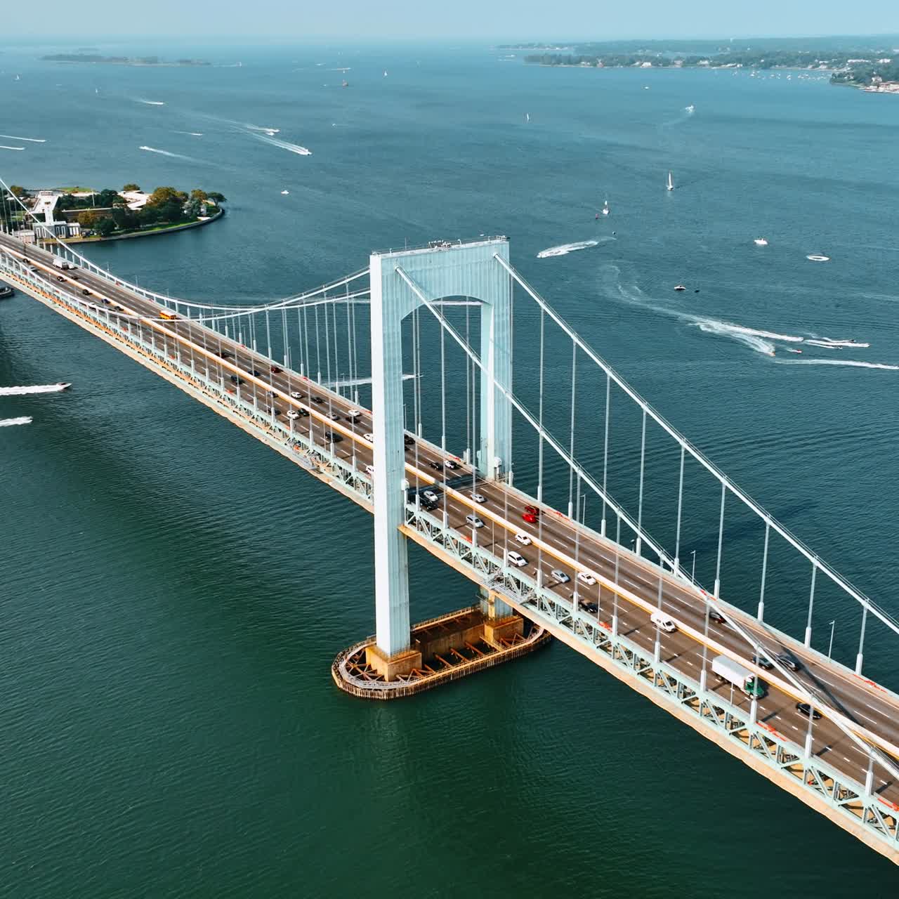 Lively traffic on the Throgs Neck Bridge on sunny day. View of the East River with multiple boats on. Aerial perspective