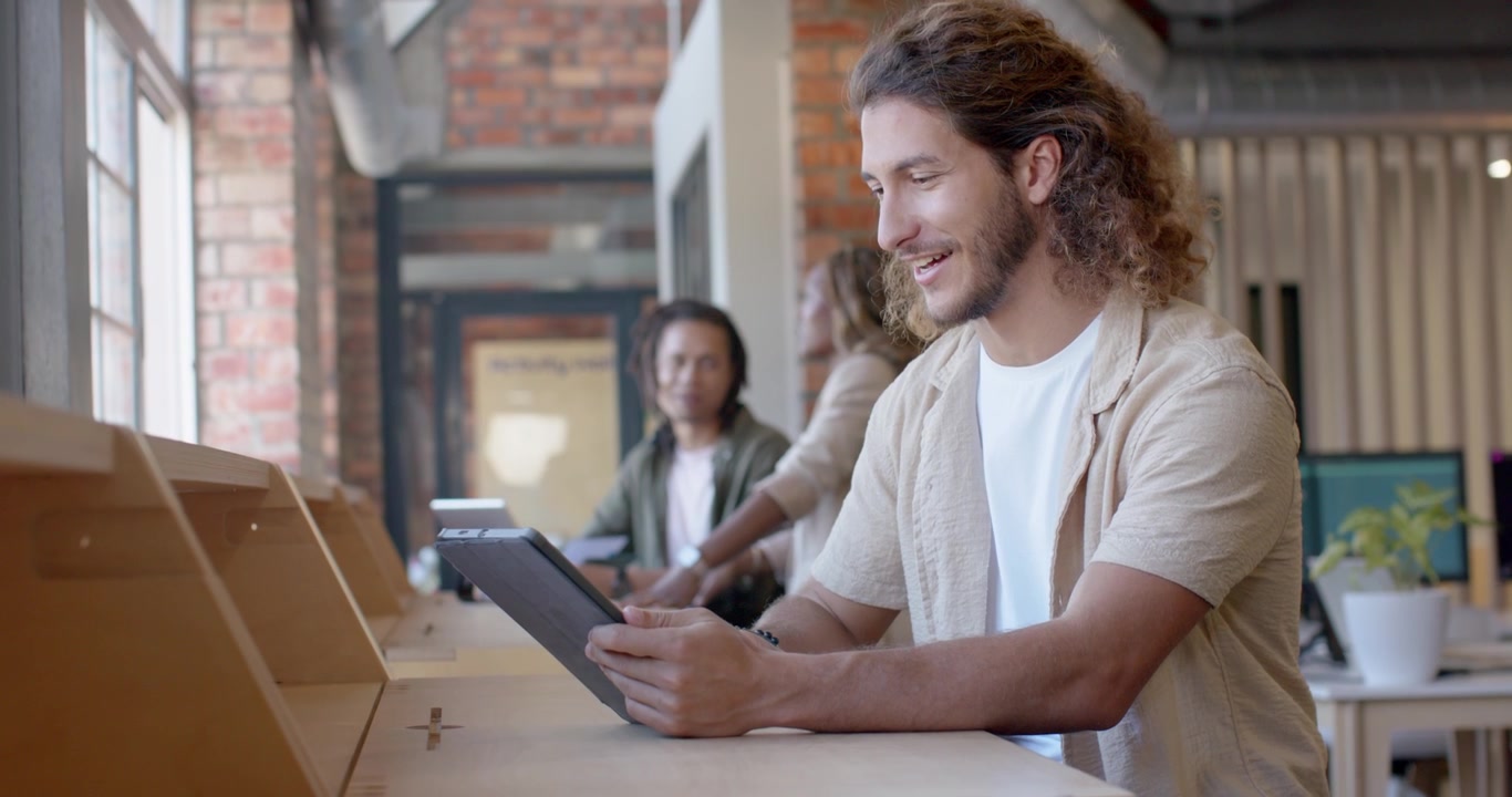Adult man tapping tablet, gesturing and speaking at long wooden counter by windows, sharing content