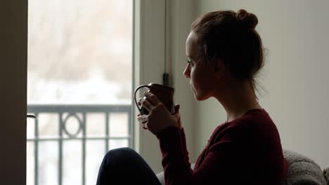 medium shot of young woman quietly watching snowflakes fall outside and enjoying hot beverage in mug