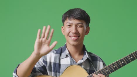 close up of asian teenager with guitar waving hand and smiling to camera on green screen background