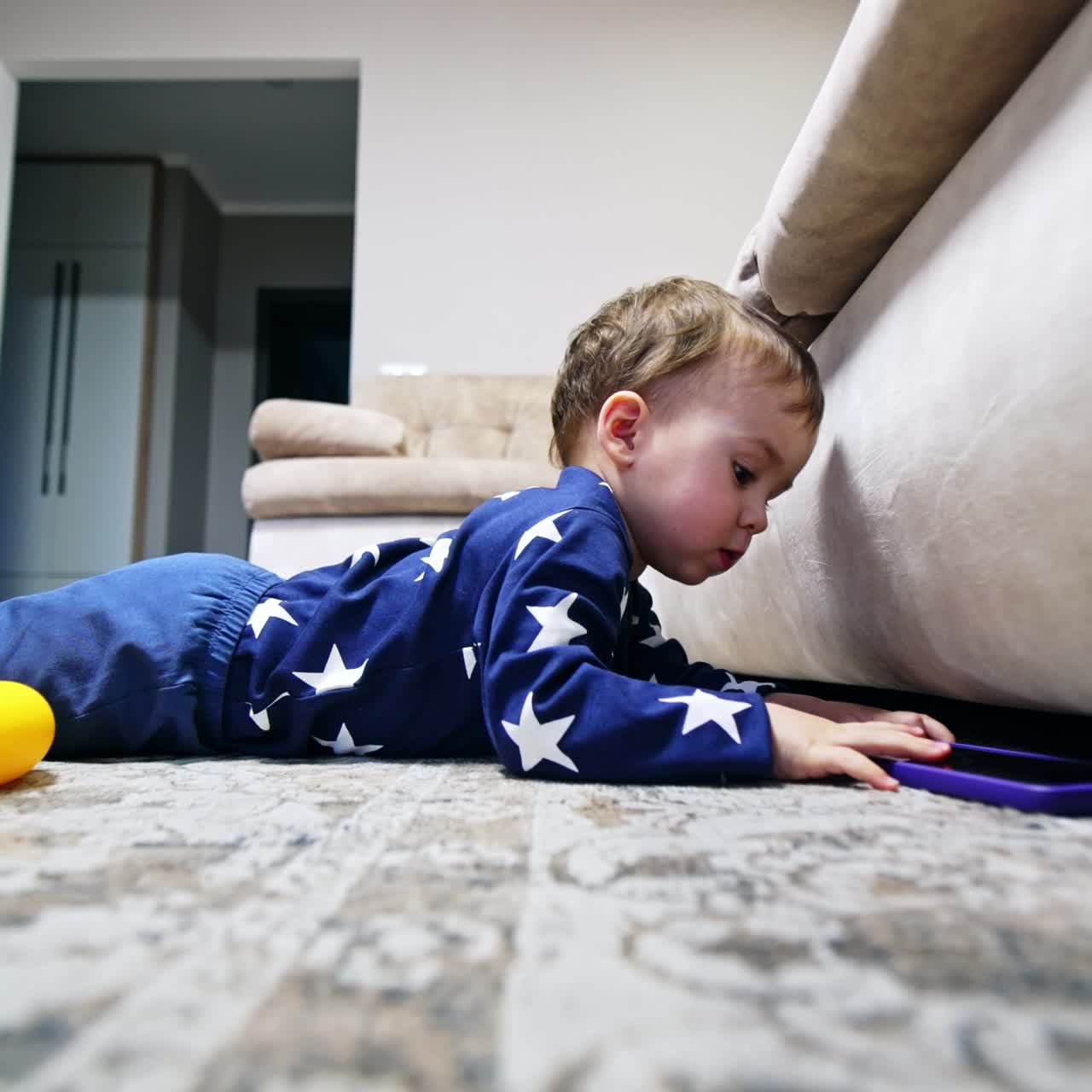 Little kid takes the phone out of the sofa. Baby boy in blue outfit lies on the floor reaching hand under the furniture