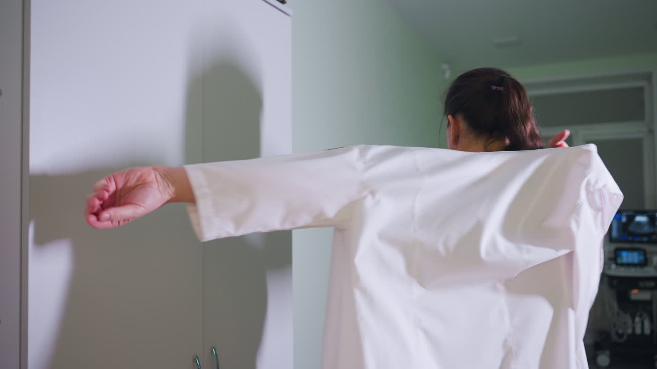 Elegant doctor stands in front of cabinet putting on work clothing while her shadow softly reflects on cabinet surface in softly lit clinic room with medical equipment visible in background