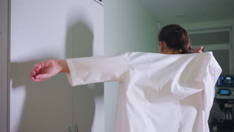 elegant doctor stands in front of cabinet putting on work clothing while her shadow softly reflects on cabinet surface in softly lit clinic room with medical equipment visible in background