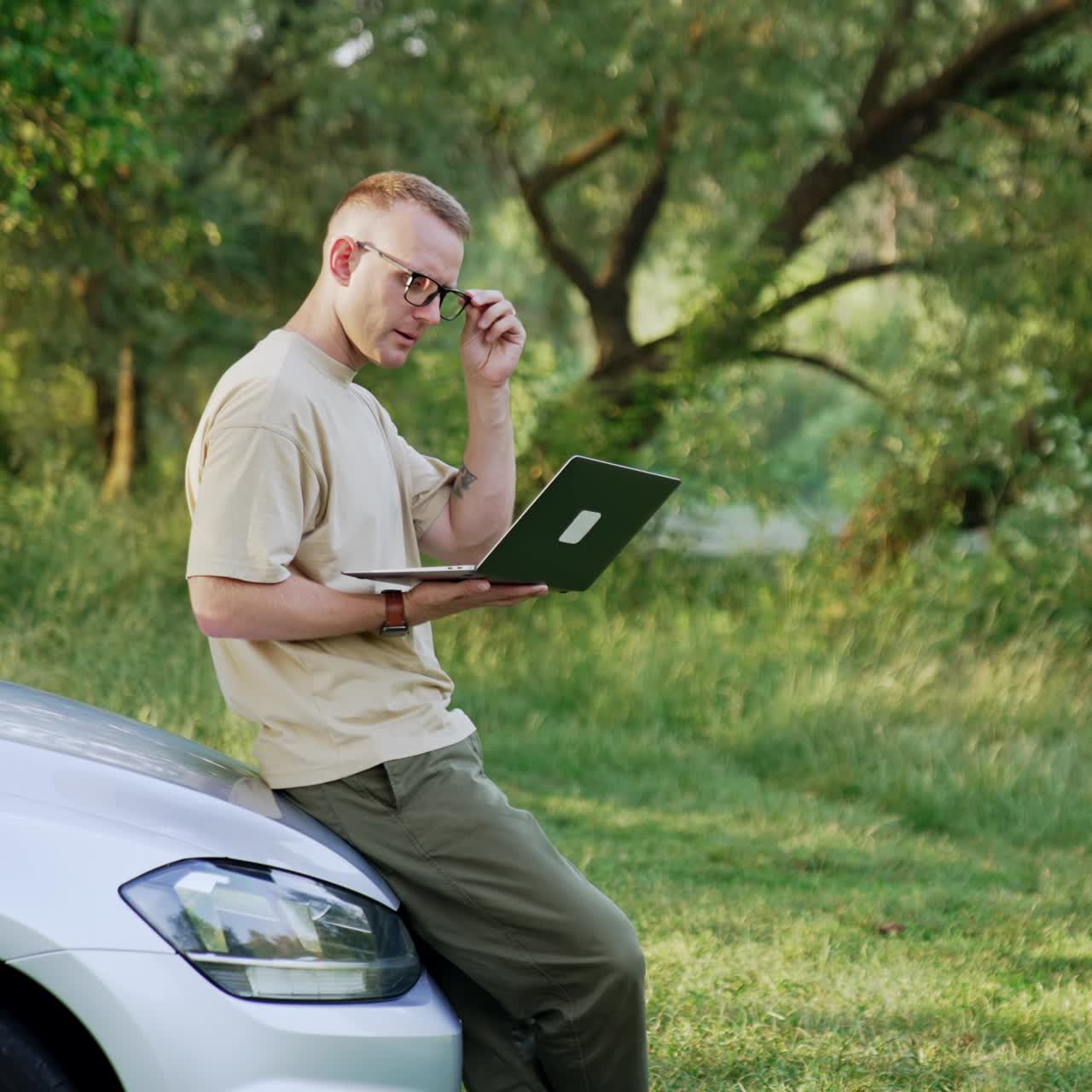 Mid-aged man stands leaning on car outdoors. Man works on laptop holding it in hands. Freelancer takes off glasses looking ahead pensively