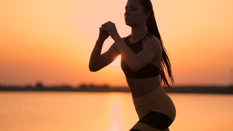 Seitenansicht-Einer-Jungen-Frau,-Die-Draußen-Kniebeugen-Macht.-Seitenansicht-Einer-Jungen-Fitness-Frau,-Die-Kniebeugen-Macht-Und-Bei-Sonnenuntergang-In-Zeitlupe-Am-See-Auf-Dem-Sand-Steht