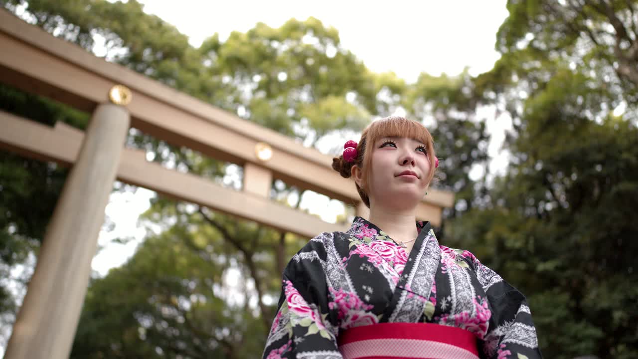 Una donna giapponese in kimono floreale sta davanti a un grande santuario con portale torii in legno.