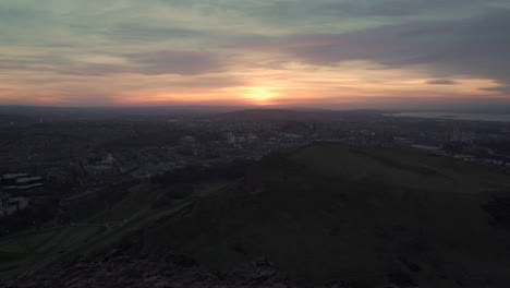 Statische-Aufnahme-Der-Stadtlandschaft-Von-Edinburgh-Vom-Berg-Aus,-Momente-Nach-Sonnenuntergang-Von-Arthurs-Seat-Aus-Mit-Wundervollem-Sonnenuntergang,-Blauem-Stundenlicht-Und-Unten-Vorbeifahrenden-Autos