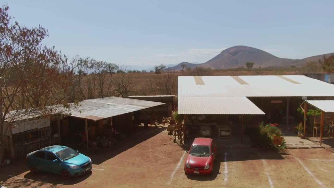 Drone pull out from rural farm buildings with parked cars and mountain landscape. Expanding aerial perspective reveals open countryside, dry vegetation and surrounding agricultural land