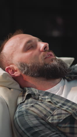 bearded man with acute heart pain lies on sofa putting hand on chest. male with high temple waits for relief after taking medications in living room closeup