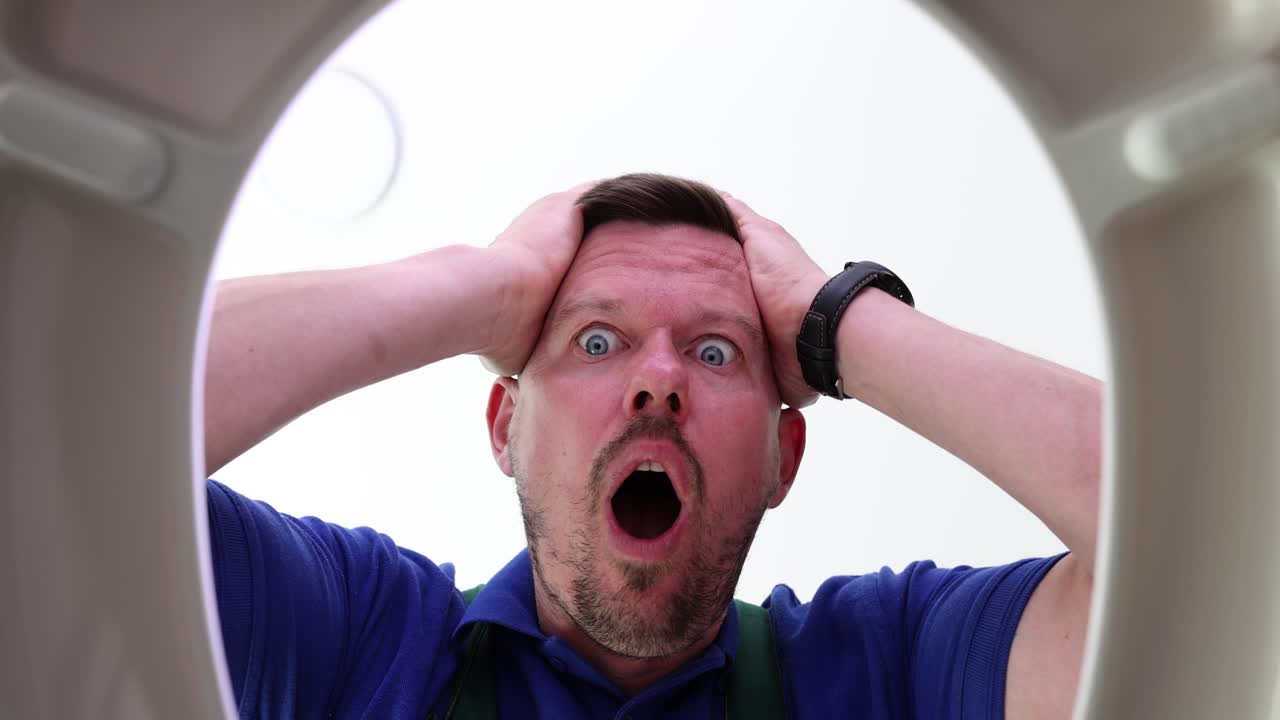 Shocked male plumber opens toilet lid putting hands on head