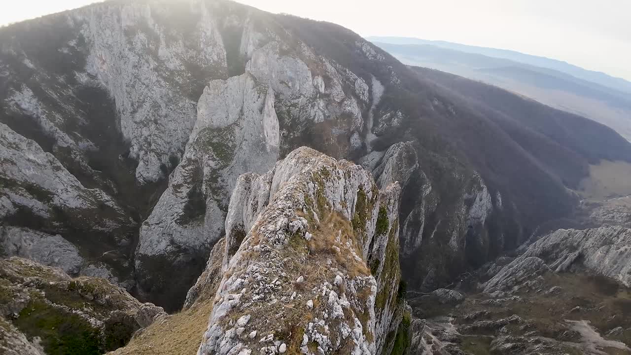 una toma de zoom aéreo que muestra un acantilado de montaña escarpado con un profundo valle debajo, enfatizando el terreno rocoso dramático y empinado