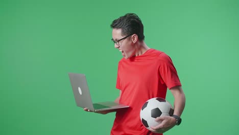 vista lateral de un hombre asiático con una pelota gritando gol alegrándose por el equipo ganador mientras anima el fútbol en una computadora portátil en el estudio de pantalla verde