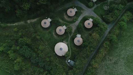 ojo de pájaro de arriba hacia abajo seguimiento aéreo hacia adelante sobre un grupo de chozas de glamping rodeadas de bosque y campo verde