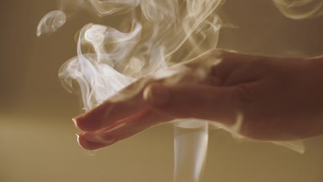 close up female hand playing with white curling fume indoors natural light slow motion. faceless woman stroking incense smoke swirling in sunlight shallow crop view. meditation relaxation calmness