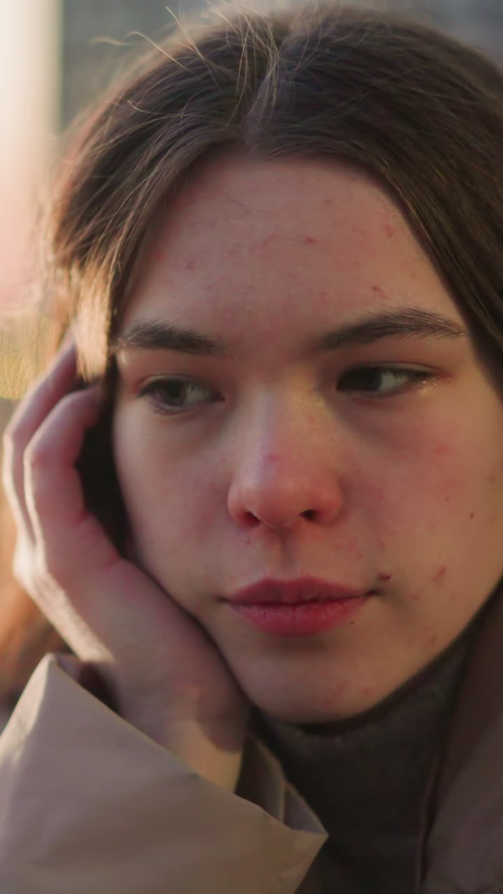A close-up shot of a girl in a peach jacket, lost in deep thought as she gently rests her cheek on her hand. A faint smile graces her face. The blurred background with evening lights