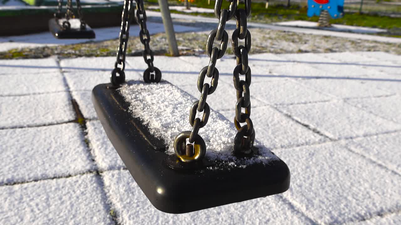 Close up view of a playground swing covered in white fluffy and fresh first snow during a winter sunny day with green grass and others playground attractions in the background