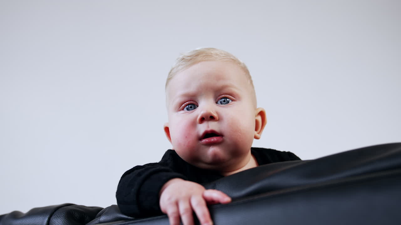 Blond grey-eyed baby boy with cute plump cheeks. Child stands on the black leather sofa looking focused down. Low angle view.
