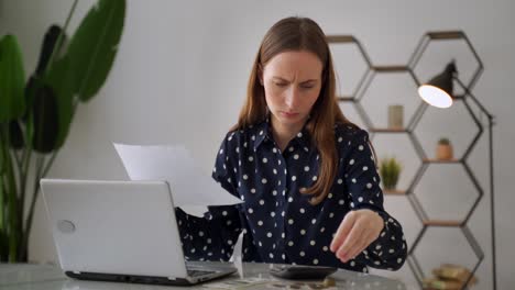 una mujer preocupada está sentada en un escritorio, usando una computadora portátil, comprobando las finanzas usando una calculadora