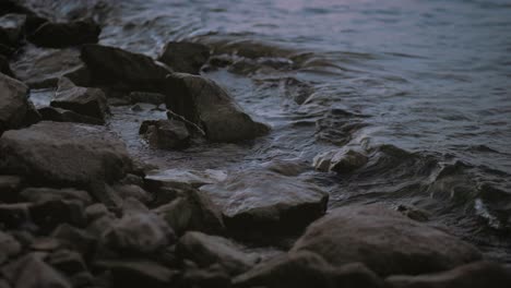 A-static-detail-shot-of-small-waves-on-the-lake-during-dusk-in-slow-motion-Summer-evening-on-the-riverside,-stones-washed-by-small-waves